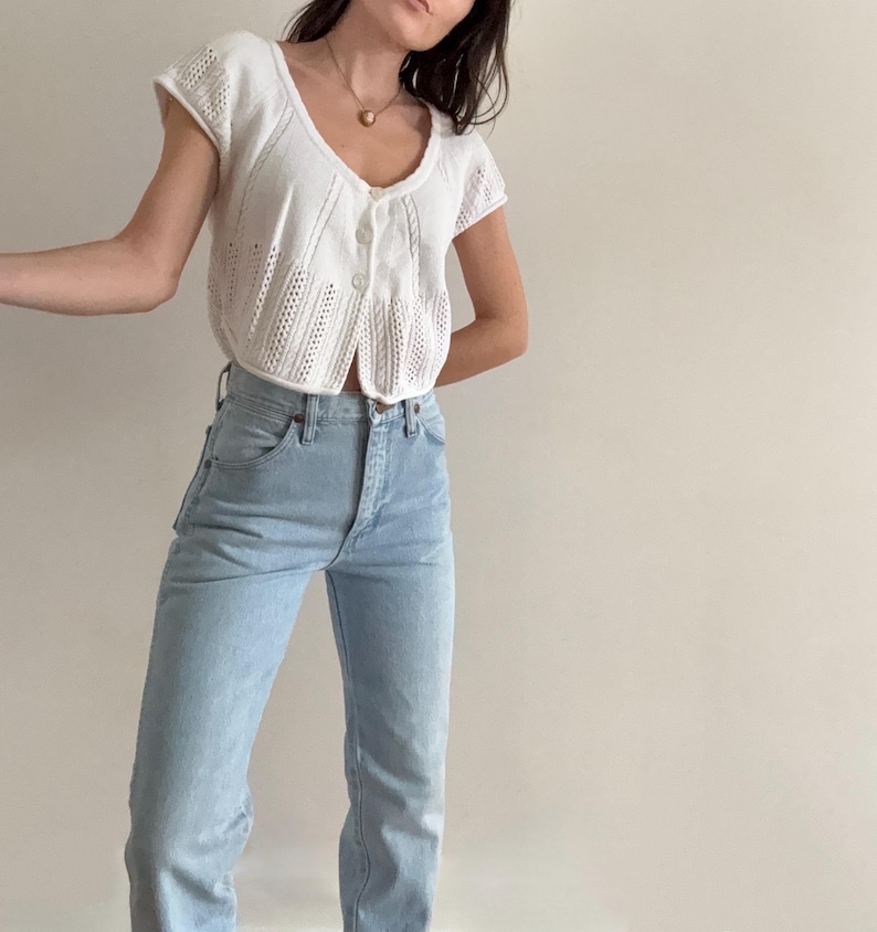 May include: A white, short-sleeved knit cardigan with a button-up front and a scalloped hemline. Paired with light blue denim jeans. The cardigan has a delicate, open-weave design. The jeans are high-waisted with a straight leg.