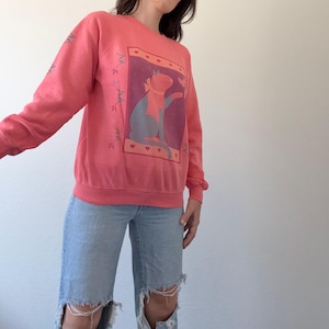 May include: A coral-coloured sweatshirt with a cat graphic in a rectangular frame with hearts. The cat is blue and orange. The sleeves have a floral design. The sweatshirt is worn with light blue ripped jeans.