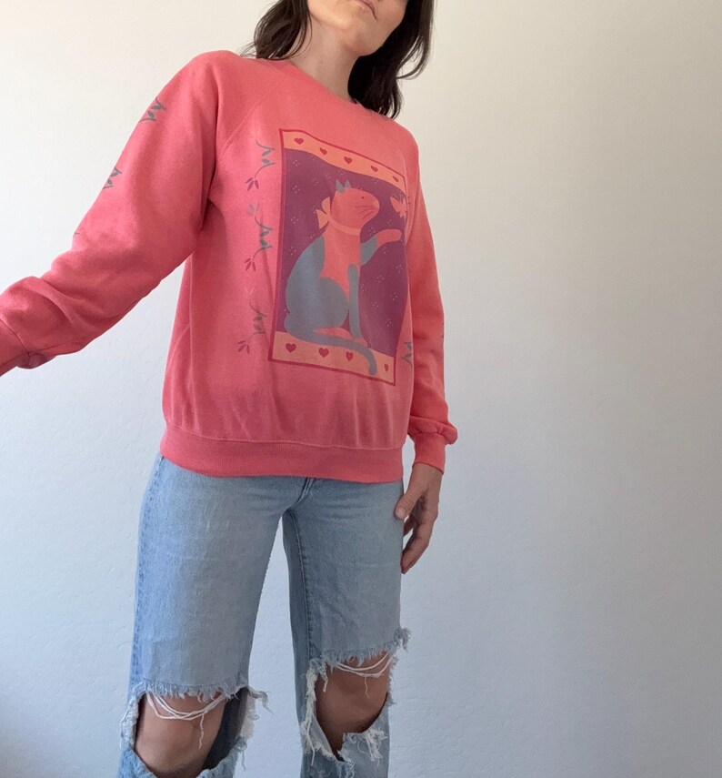 May include: A coral-coloured sweatshirt with a cat graphic in a rectangular frame with hearts. The cat is blue and orange. The sleeves have a floral design. The sweatshirt is worn with light blue ripped jeans.