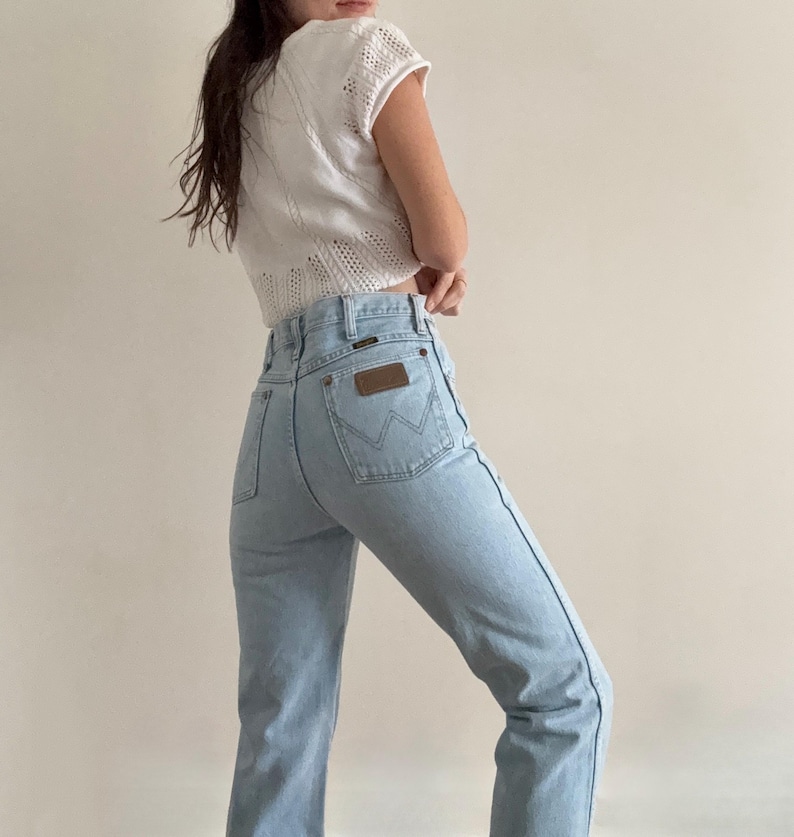May include: Light blue denim jeans with a high waist and a brown leather patch on the back pocket. The jeans are paired with a white, open-knit top. The Wrangler logo is visible on the back pocket.