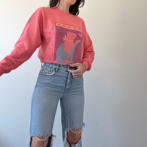 May include: A coral-coloured sweatshirt with a cat graphic, set against a purple background with hearts. The sleeves have a decorative design. The sweatshirt is worn with light blue ripped jeans. The cat graphic has a bow.