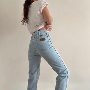 May include: Light blue denim jeans with a high waist and a brown leather patch on the back pocket. The jeans have a classic five-pocket design and a straight leg. The model is wearing a white, open-knit top.