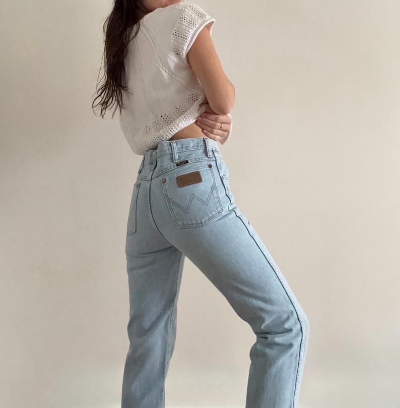 May include: Light blue denim jeans with a high waist and a brown leather patch on the back pocket. The jeans have a classic five-pocket design and a straight leg. The model is wearing a white, open-knit top.