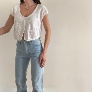 May include: A white, short-sleeved cardigan with a V-neck and button closure. The cardigan has a textured knit design. Paired with light blue denim jeans.