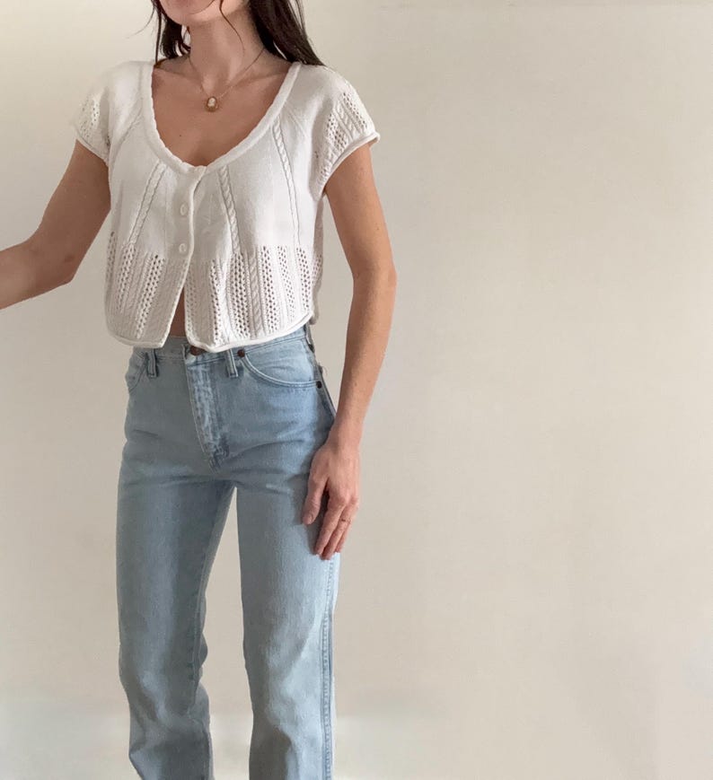 May include: A white, short-sleeved cardigan with a V-neck and button closure. The cardigan has a textured knit design. Paired with light blue denim jeans.