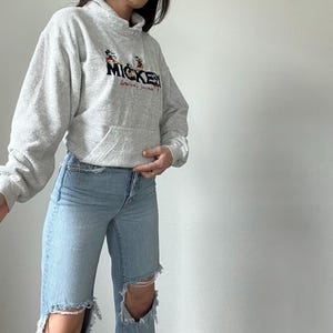 May include: A gray hooded sweatshirt with a Mickey Mouse graphic and the text "Mickey, America's Favorite" printed on the front. The sweatshirt is paired with light blue jeans with distressed details.