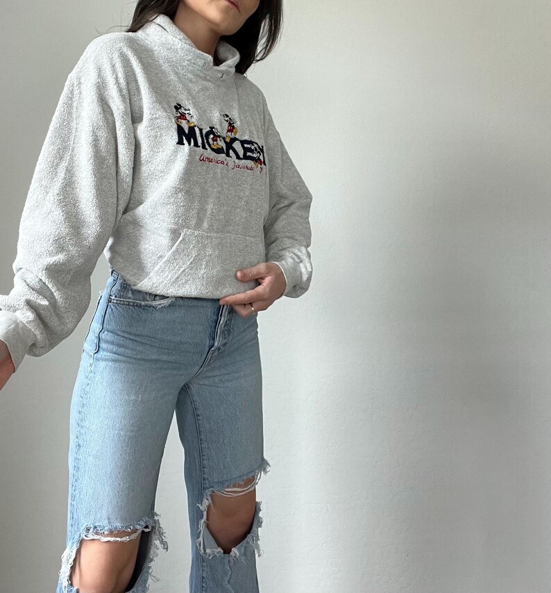 May include: A gray hooded sweatshirt with a Mickey Mouse graphic and the text "Mickey, America's Favorite" printed on the front. The sweatshirt is paired with light blue jeans with distressed details.