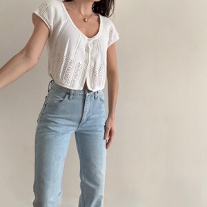 May include: A white, short-sleeved knit top with a button-up front and a scoop neckline. The top is paired with light blue high-waisted jeans. The outfit is simple and classic, suitable for casual wear.