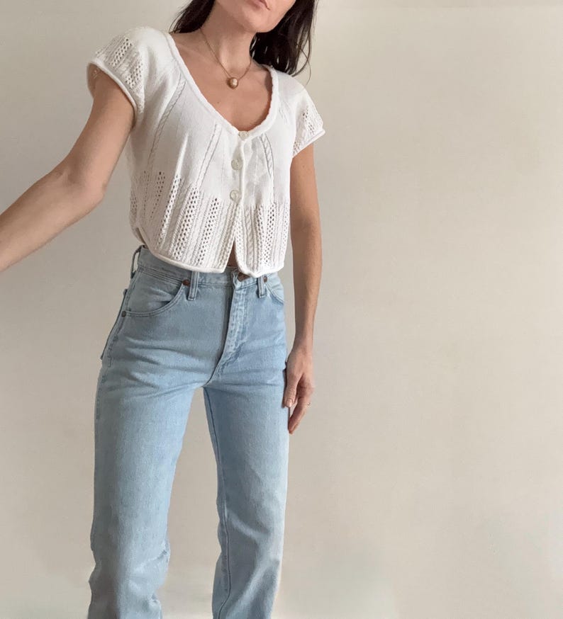 May include: A white, short-sleeved knit top with a button-up front and a scoop neckline. The top is paired with light blue high-waisted jeans. The outfit is simple and classic, suitable for casual wear.