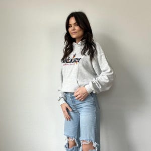 May include: A gray hooded sweatshirt with a Mickey Mouse graphic and the text "Mickey Mouse" in red and black lettering. The model is wearing light blue distressed jeans.