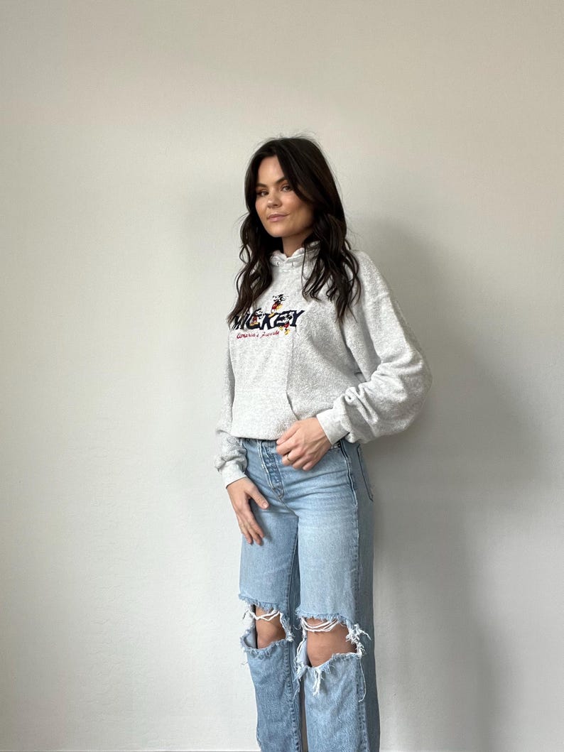 May include: A gray hooded sweatshirt with a Mickey Mouse graphic and the text "Mickey Mouse" in red and black lettering. The model is wearing light blue distressed jeans.