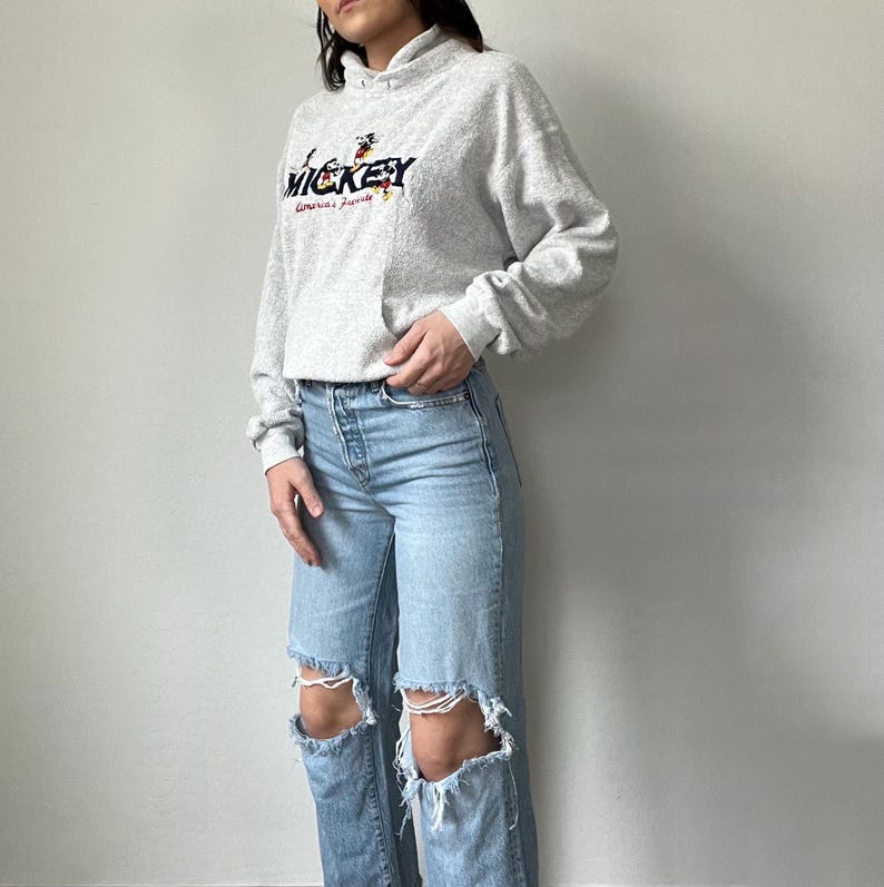 May include: A gray sweatshirt with a Mickey Mouse graphic and the text "Mickey America's Favorite" on the front. The sweatshirt has a hood and a pouch pocket. The model is wearing blue jeans with distressed details.