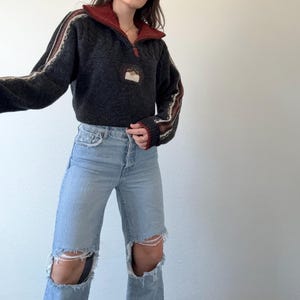 Puede incluir: Un su&eacute;ter gris oscuro con cuello y cremallera burdeos. Las mangas tienen rayas crema, marr&oacute;n y burdeos. El su&eacute;ter tiene un peque&ntilde;o parche en el pecho. La modelo lleva vaqueros rotos de color azul claro.