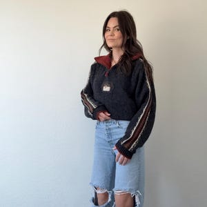 Puede incluir: Un su&eacute;ter gris oscuro con cuello rojo y cuello con cremallera. Las mangas tienen rayas marrones y blancas. El su&eacute;ter tiene un peque&ntilde;o parche en el pecho. La persona lleva vaqueros rotos de color azul claro.