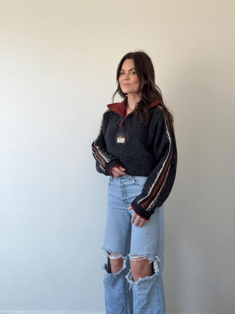 Puede incluir: Un su&eacute;ter gris oscuro con cuello rojo y cuello con cremallera. Las mangas tienen rayas marrones y blancas. El su&eacute;ter tiene un peque&ntilde;o parche en el pecho. La persona lleva vaqueros rotos de color azul claro.