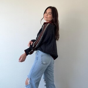 Puede incluir: Un su&eacute;ter gris oscuro con rayas estampadas en las mangas y el cuello. El su&eacute;ter se combina con unos vaqueros azules claros rotos. El conjunto es informal y c&oacute;modo.