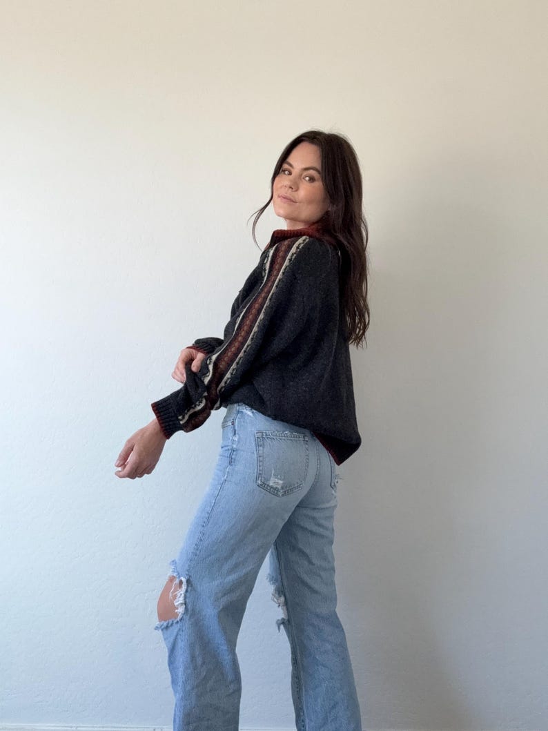 Puede incluir: Un su&eacute;ter gris oscuro con rayas estampadas en las mangas y el cuello. El su&eacute;ter se combina con unos vaqueros azules claros rotos. El conjunto es informal y c&oacute;modo.