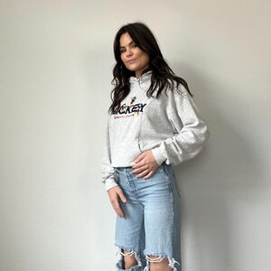 May include: A gray hooded sweatshirt with a Mickey Mouse graphic and the text "Mickey" in colorful lettering. The sweatshirt is worn by a person with long brown hair and is paired with light blue jeans with distressed details.