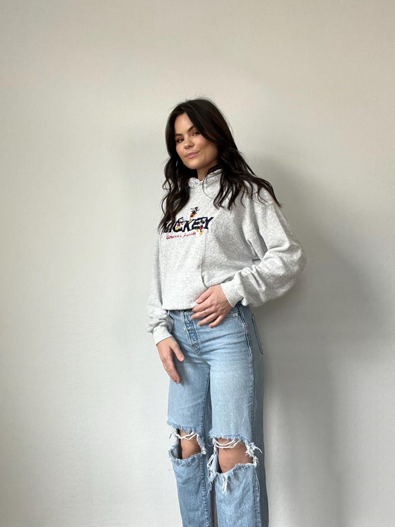 May include: A gray hooded sweatshirt with a Mickey Mouse graphic and the text "Mickey" in colorful lettering. The sweatshirt is worn by a person with long brown hair and is paired with light blue jeans with distressed details.