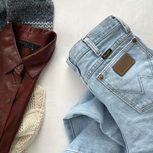 May include: An assortment of clothing items including a light blue denim Wrangler jeans, a brown leather shirt with the "Express" label, and a patterned sweater. A cream-colored sweater is also visible.