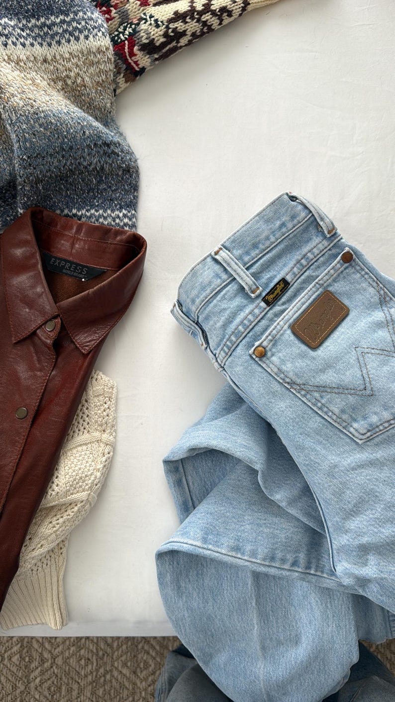 May include: An assortment of clothing items including a light blue denim Wrangler jeans, a brown leather shirt with the "Express" label, and a patterned sweater. A cream-colored sweater is also visible.
