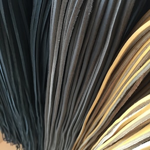 Puede incluir: Una colección de tiras de cuero largas y delgadas dispuestas en filas verticales. Las tiras están agrupadas por color, presentando negro profundo, gris carbón oscuro y un tono beige o bronceado claro. Textura mate y uniforme.
