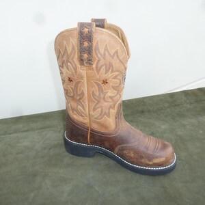 May include: A tan and dark brown leather western boot featuring a square toe, a black rubber sole, and decorative stitching on the shaft. The upper shaft includes a dark brown pull tab with star cutouts and small embroidered star details.