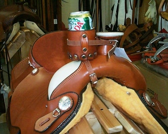 May include: A brown leather western saddle featuring a cylindrical leather beverage holder attached to the pommel with metal rivets and a buckle strap. The saddle includes silver-toned decorative conchos and a cream-colored fleece underside.