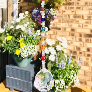 May include: A crystal suncatcher with a rainbow of coloured beads hanging from a clear crystal ball. The suncatcher is hanging in front of a brick wall and green plants.