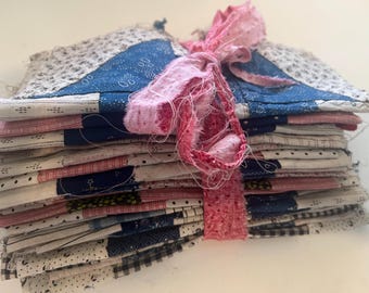 May include: A stack of handmade fabric squares, featuring a patchwork design in blue, white, and red hues. The squares are tied together with a pink, frayed ribbon. The overall aesthetic suggests a vintage or handcrafted theme.