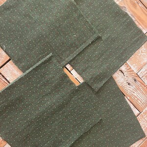 May include: A collection of six square, dark green fabric napkins with a subtle dotted pattern in a lighter green. The napkins are arranged on a light brown wooden surface, showcasing their texture and design. Ideal for home decor or dining.