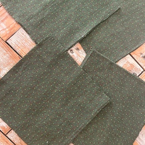 May include: Four dark green fabric napkins with a subtle dotted pattern in a lighter green. The napkins are arranged on a wooden surface, showcasing their texture and design. The fabric appears to be a woven material.