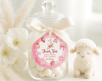 May include: A clear glass jar filled with white candies, adorned with a pink tag and a cream ribbon. The tag reads "Thank You! Berry Much For Coming!" and features strawberry illustrations. A small lamb figurine sits beside the jar.