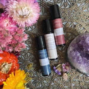 May include: Three aromatherapy roller bottles with labels that read "Peace", "Happiness", and "Bliss". The bottles are on a gold metal surface with purple amethyst crystals and flowers.