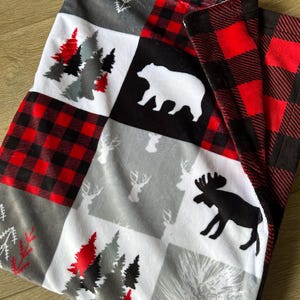 Peut inclure: Une couverture patchwork avec des motifs de forêt et d'animaux sauvages. La couverture comprend des carreaux à carreaux rouges et noirs, des silhouettes d'ours blancs sur fond noir et des motifs de cerfs et d'arbres gris. La palette de couleurs est rouge, noir, blanc et gris.