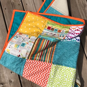 Peut inclure: Une courtepointe patchwork colorée sur le thème de la science. La courtepointe présente des carrés de tissus turquoise, jaunes, oranges et blancs avec divers motifs, notamment des pois, des chevrons et du texte et des graphiques liés à la science. La courtepointe est bordée d'une bordure orange.