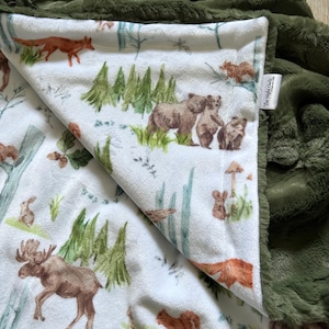 May include: A soft, plush blanket featuring a white side with a watercolour-style forest print of bears, a moose, a fox, a squirrel, and pine trees. The reverse side is a solid olive green faux fur. A white fabric tag reads The Night Owls.