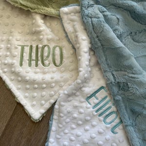 May include: A selection of baby blankets in various colours, including green and blue. One white blanket is embroidered with the name "THEO" in green, and another with "Elliot" in blue. The blankets have a soft, textured finish.