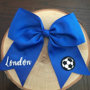 May include: A royal blue hair bow with a white soccer ball and the name "London" embroidered on it.