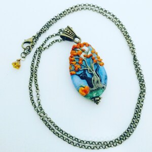 May include: A black cat sits in a tree with orange leaves. The tree is on a blue and white oval pendant with a green and brown base. The pendant is attached to a bronze chain necklace.