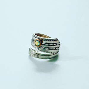 May include: A silver ring with a decorative design and a small yellow and blue stone in the center.