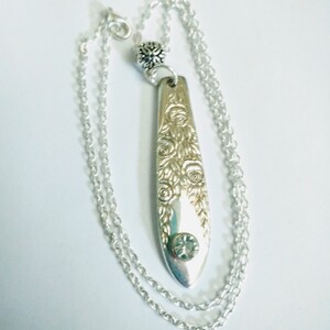 May include: A silver necklace with a long, thin pendant featuring a floral design and a clear rhinestone. The pendant is attached to a silver chain.