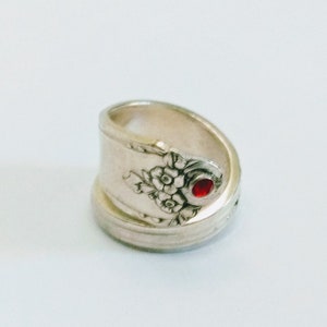 May include: A silver ring made from a repurposed spoon with a red gemstone set in the center. The spoon has a floral design.