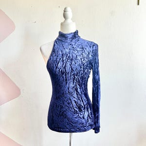 May include: A dark blue crushed velvet top with a high neck and one long sleeve. The top is displayed on a white mannequin. The fabric has a textured, wrinkled appearance.
