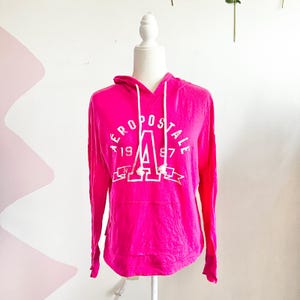 Könnte beinhalten: Ein leuchtend pinkfarbener Kapuzenpullover mit dem weißen Schriftzug "AEROPOSTALE 1987". Der Pullover hat eine Kapuze mit Kordelzug und eine Vordertasche. Das langärmlige Oberteil wird auf einer Schaufenquin präsentiert.