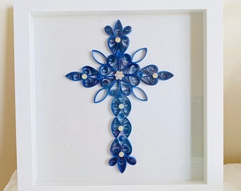 May include: A blue paper quilling cross with white embellishments framed in a white frame.