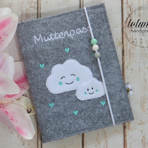 Könnte beinhalten: Grauer Filzbuchumschlag mit weißer Stickerei, die "Mutterpass" sagt. Der Umschlag zeigt zwei weiße Filzwolken mit lächelnden Gesichtern und türkisfarbenen Regentropfen. Ein weißes Band mit türkisfarbenen und weißen Perlen ist am Umschlag befestigt.