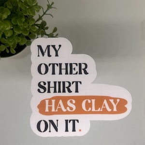May include: White sticker with black text that reads "My other shirt has clay on it." The words "has clay" are in a brown rectangle.