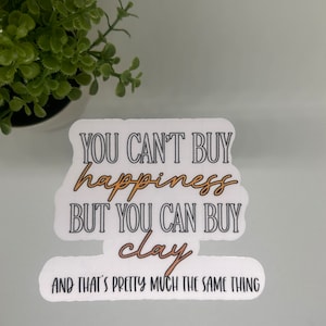 May include: White sticker with black text that reads "You can't buy happiness but you can buy clay and that's pretty much the same thing". The word "clay" is in orange.