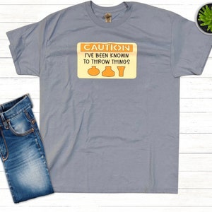 Puede incluir: Una camiseta gris jaspeada con una señal de advertencia amarilla que dice "Precaución He sido conocido por tirar cosas" con tres símbolos de cerámica naranja.